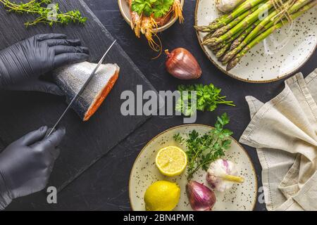 Hände in schwarzen Handschuhen schneiden Forellenfisch auf schwarzen Stein Schneidebrett umgeben Kräuter, Zwiebel, Knoblauch, Spargel, Garnelen, Garnelen in Keramikplatte. Schwarzer CO Stockfoto