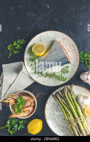 Forellenfisch umgab Petersilie, Zitrone, Garnelen, Garnelen, Spargel in Keramikplatten. Tischfläche aus schwarzem Beton. Gesunde Meeresfrüchte Hintergrund. Stockfoto