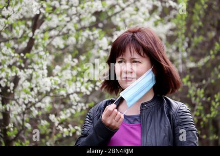 Freudige Frau Entfernen medizinische Schutzmaske im Frühlingsgarten auf Kirschblüten Hintergrund. Konzept des Genußens der Blumen Geruch, Ende der Quarantäne Stockfoto