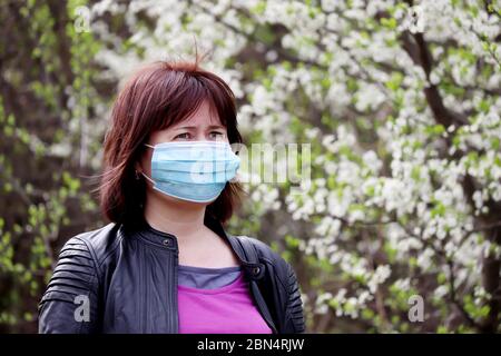 Frau in medizinischer Schutzmaske in einem Frühlingsgarten auf Kirschblütengrund. Konzept der Quarantäne während der Covid-19 Coronavirus-Pandemie Stockfoto