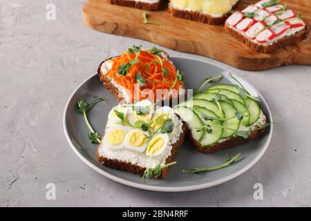 Drei Sandwiches auf Toast mit Frischkäse, Karotten, Gurken und Wachteleiern mit Mikrogreens Erbsen auf einem Teller auf grauem Beton Hintergrund dekoriert Stockfoto