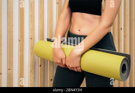 Nahaufnahme einer jungen sportlichen Frau, die mit einer grünen Yogamatte auf der weißen und hölzernen Wand steht. Ruhe nach Yoga-Übungen, gemütliche und soziale Atmosphäre in Stockfoto