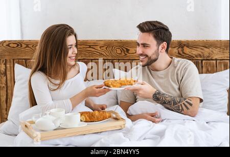Romantisches Frühstück für zwei Personen. Mädchen gibt Croissant zu Mann, im Bett Stockfoto