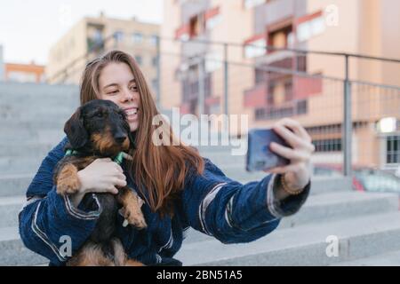 Alternative hübsche junge Erwachsene Frau mit blauer Jacke macht ein Selfie-Foto mit ihrem Hund im Freien in der Stadt Stockfoto