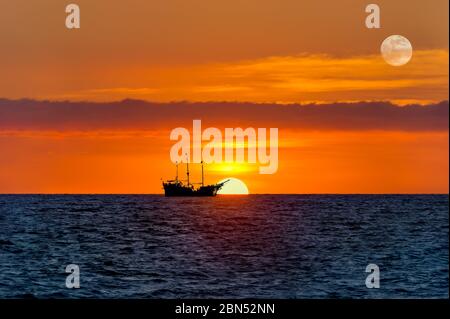 Ein altes Holzschiff sitzt auf See, während die Sonne am Ozeanhorizont untergeht und der Mond in den Himmel steigt Stockfoto