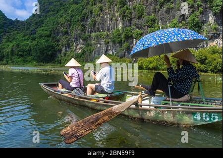 Ninh Binh Provinz, Vietnam - 22. Oktober 2019 Menschen in traditionellen vietnamesischen Hüten im Boot entlang des Flusses. Zwei Touristen schauen sich die Telefone an. Tam Coc Boat Ri Stockfoto