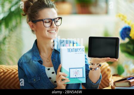 Lächelnd stilvolle Mittelalterhausfrau in Jeans-Shirt in modernen Hause in sonnigen Tag zeigt Lehrbuch und Tablet-PC leeren Bildschirm. Stockfoto