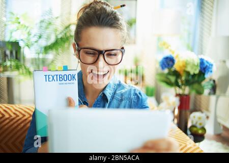 Lächelnde moderne Hausfrau in Jeans-Hemd zeigt Lehrbuch während einer Videokonferenz mit einem Lehrer auf einem Tablet-PC im modernen Haus an sonnigen Tag. Stockfoto