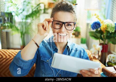 Portrait der glücklichen jungen Frau in Jeans-Shirt mit weißen Kopfhörern und Tablet-PC Online studieren im modernen Wohnzimmer an sonnigen Tag. Stockfoto