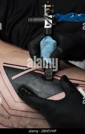 Professioneller Tattoo-Künstler macht Tattoo am Mannbein. Prozess. Führt schwarze Tinte mit der Nadel von der Tattoo-Maschine in die Haut ein. Nahaufnahme der Tattoo-Maschine. Tätowierung. Erstellen eines Bildes auf dem Bein. Stockfoto