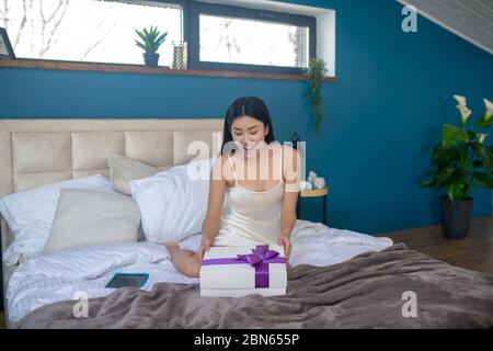 Junge Brünette in Nachtschleidchen auf dem Bett sitzen, lächelnd, Blick auf Geschenk-Box mit lila Band Stockfoto
