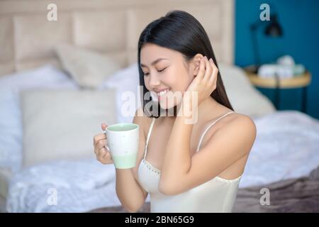 Junge Brünette im Nachtschleifkleid sitzt auf dem Bett, hält weiße Tasse, trinken, lächeln, mit einer Hand über das Haar laufen Stockfoto