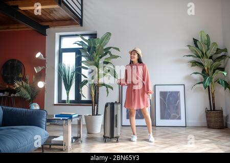 Aufgeregt dunkelhaarige Frau in Strohhut hält ihren Koffer Griff, lächelnd Stockfoto