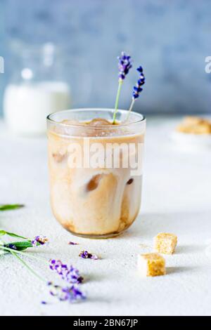 Glas Kaffee mit Eis, Milch und Lavendelblüten auf einem hellen Hintergrund. Kaltes Sommergetränk. Stockfoto