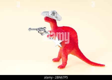 Nahaufnahme eines kleinen roten Spielzeugdinosauriers, der einen Cowboy-Hut trägt, einen Revolver im Arm hält und einen Sheriff-Stern auf neutralem Hintergrund hat. Stockfoto