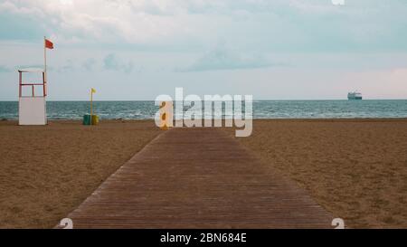 Verlassene Strand mit Holzplanken durch den Sturm. Mit einer roten Flagge Warnung vor Gefahr. Im Hintergrund tritt ein Kreuzfahrtschiff zurück. Stockfoto