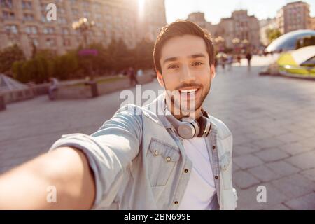 Selfie-Manie! Aufgeregt junger Kerl macht Selfie auf einer Kamera. Er trägt lässige trendige Kleidung und große moderne Kopfhörer, auf einem Spaziergang in der Frühlingsstadt Stockfoto