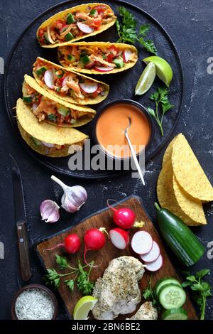 Büffel Hühnermast Taco Muscheln mit frischem Gemüse und Gemüse auf einem schwarzen Teller mit Limette und Büffelsauce mit Zutaten auf einem Beton t geladen Stockfoto