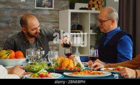 Junger Mann, der seinen Schwiegervater beim Familienessen mit Rotwein bedient. Leckeres Essen. Stockfoto