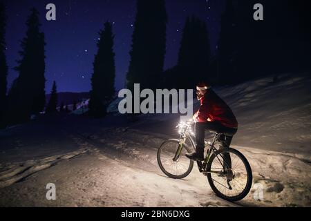 Mann im roten Mantel mit Fahrrad und Weihnachten hat im Winter verschneite Wald in den Bergen unter Nachthimmel mit Sternen Stockfoto