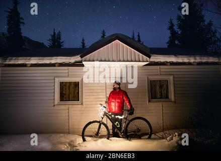 Mann im roten Mantel mit Fahrrad mit Weihnachtsbeleuchtung in der Nähe von kleines Haus im Winter verschneite Wald in die Berge unter dem Nachthimmel mit Sternen Stockfoto