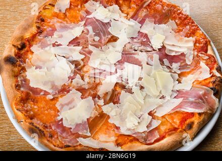 Italienische Pizza Ofen gebacken traditionell mit Tomatensauce, Mozzarella, Prosciutto, Rucola, Parmesan Späne. Einfache braune hölzerne Restaurant Tisch-Setti Stockfoto