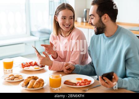 Porträt von jungen fröhlichen Paar mit Handys und Lachen beim Frühstück in der gemütlichen Küche zu Hause Stockfoto