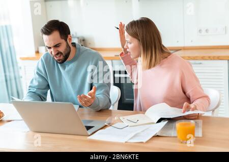Portrait des jungen wütenden Paares, das mit Laptop und Dokumenten in der gemütlichen Küche zu Hause arbeitet Stockfoto