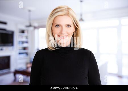 Porträt der schönen blonden Frau, die Kamera anschaut und lächelt, während sie im Wohnbereich zu Hause steht. Stockfoto
