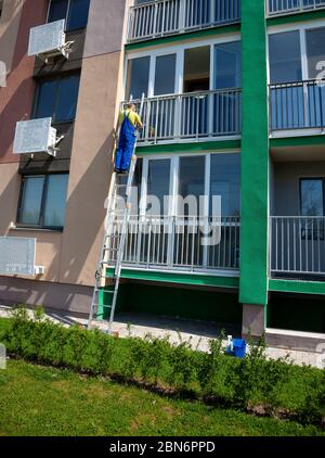 Ein Mann wäscht die Fenster auf der Straße, die auf der Treppe stehen. Reinigungsfirma. Arbeiter in blauen Overalls wäscht ein Fenster einer mehrstöckigen Wohnung Stockfoto