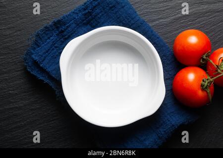 Draufsicht leere leere Mock-up weiße Keramik gebackene Schüssel mit blauen Leinen Serviette auf schwarzem Schieferstein Brett Stockfoto