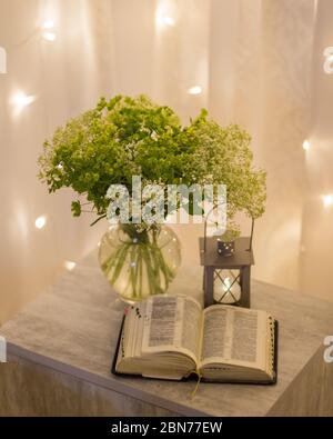 Komposition: Eine offene Bibel, ein Blumenstrauß mit Wildblumen in einer Vase und eine dekorative Taschenlampe mit einer Kerze auf dem Nachttisch vor dem Hintergrund von b Stockfoto