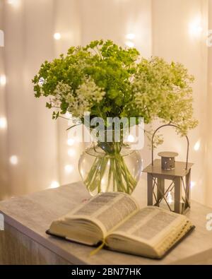 Komposition: Eine offene Bibel, ein Blumenstrauß mit Wildblumen in einer Vase und eine dekorative Taschenlampe mit einer Kerze auf dem Nachttisch vor dem Hintergrund von b Stockfoto