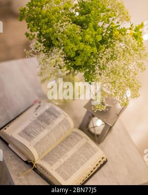 Komposition: Eine offene Bibel, ein Blumenstrauß mit Wildblumen in einer Vase und eine dekorative Taschenlampe mit einer Kerze auf dem Nachttisch vor dem Hintergrund von b Stockfoto