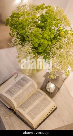 Komposition: Eine offene Bibel, ein Blumenstrauß mit Wildblumen in einer Vase und eine dekorative Taschenlampe mit einer Kerze auf dem Nachttisch vor dem Hintergrund von b Stockfoto