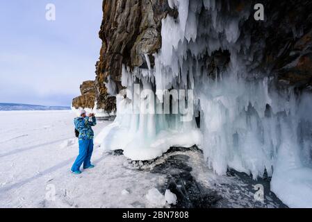 Baikalsee, Russland - 10. März 2020: Frau in blauer Jacke und weißem Hut fotografiert eisige Felsen mit Eiszapfen am Wintersee Baikalsee Stockfoto