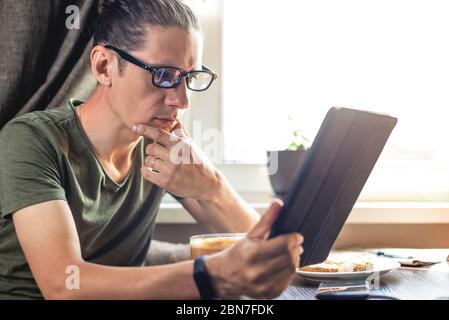 Ein junger Mann im Internet surfen und Videos mit einem Tablet am Fenster in der Wohnung. Konzept des Verbringen des Morgens zu Hause Stockfoto