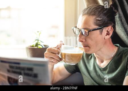 Ein junger, schöner Mann liest interessante heiße Nachrichten in einer Zeitung und trinkt eine Tasse Cappuccino-Kaffee am Fenster. Konzept der Ausgaben t Stockfoto
