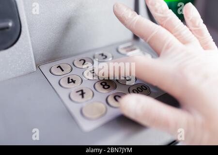 Nahaufnahme der Hand, die einen medizinischen Schutzhandschuh trägt und den PIN-Code der Kreditkarte am geldautomaten eingibt. Coronavirus-Prävention. Stockfoto