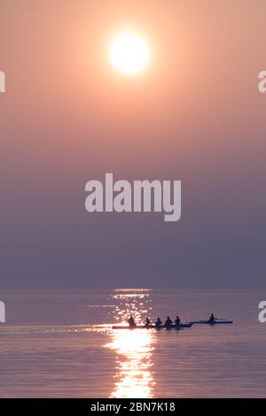 Ruderteam Training mit Trainer über dem schimmernden See bei Sonnenuntergang Stockfoto