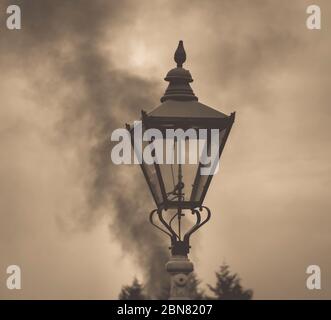 Moody, Sepia Nahaufnahme von altmodischen, Vintage Gas Licht Laterne isoliert am historischen Bahnhof mit Dampfmaschine Rauch steigt in der Luft hinter. Stockfoto