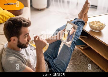 Entspannter junger bärtiger Mann, der während der Quarantäne auf dem Boden sitzt und Bier trinkt, während er sich online mit einem Freund unterhält Stockfoto