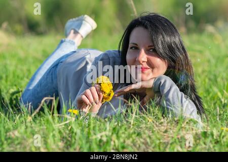 Ein schönes Mädchen, eine Brünette, liegt auf einer Blumenwiese auf dem Gras und sortiert durch gelben Löwenzahn Stockfoto