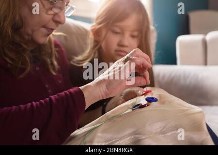 Die liebevolle Großmutter lehrt ihre kleine Enkelin, wie man Knöpfe näht Stockfoto