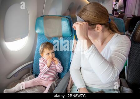 Bequemer Flug mit Kind. Niedliches kleines Kleinkind und Mama sitzen auf dem Flugzeug in Stühlen und schauen sich gegenseitig an. Mama hebt eine Schlafmaske Stockfoto