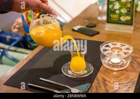 Die Hand des Menschen gießt Sanddorn Honig Tee aus Glas Teekanne in eine Tasse Glas. Nahaufnahme, Weichheit, Hintergrund verwischen Stockfoto