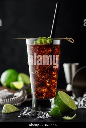 Cuba Libre Cocktail in Longballglas mit Eis und Limettenschale auf Bambusstab mit Stroh und frischen Limetten auf schwarzem Tischhintergrund. Jigger, Shaker und Stockfoto