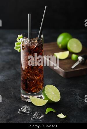 Cuba Libre Cocktail in Longdrinkglas mit Eis und Limettenschale mit Stroh und frischen Limetten auf schwarzem Tischhintergrund. Stockfoto