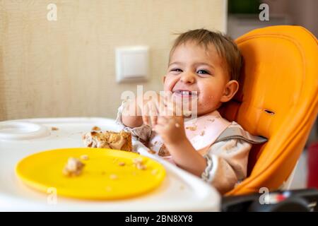 Niedliches Baby sitzt in einem orangefarbenen Babystuhl mit einem Tisch mit schmutzigen Händen und Gesicht, schaut in der Kamera und lacht. Auf dem Tisch sind Krümel und ein orangefarbener Teller Stockfoto