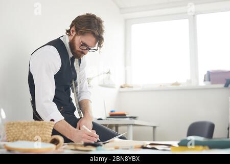 Seriöse Designer sitzen an seinem Arbeitsplatz und Notizen in Dokumenten während der Arbeit in der Werkstatt Stockfoto